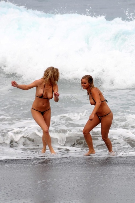 Marry Queen and her lesbian lover remove micro bikinis while the surf comes in